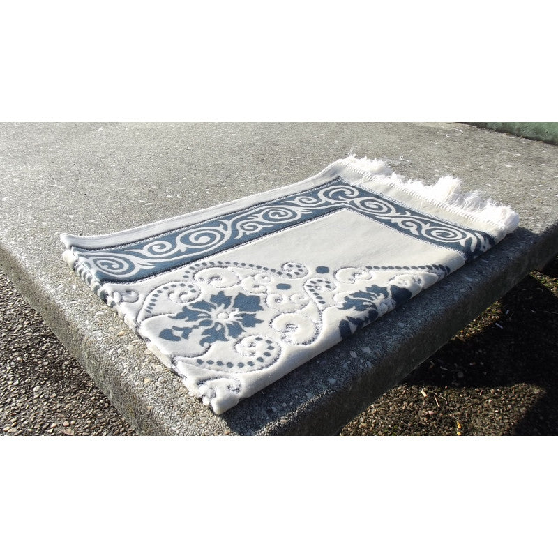 Tapis de prière Sana Edition avec fond blanc, motif fleurs, couleur bleu ardoise, disposé sur un sol.